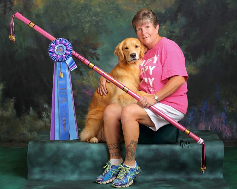 The same lady sitting beside two-year-old golden retriever, Pink in setting with a mottled green and black background and holding pink-colored PVC autograph bar with large rosette award ribbon hanging from one end-setting in a professional portrait